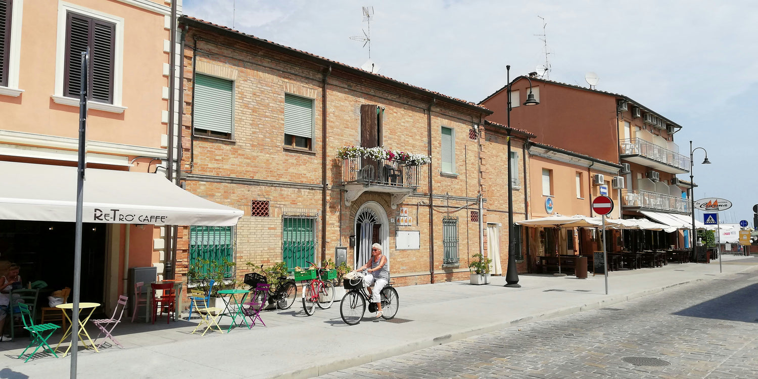 A quaint street scene in a small town, featuring a woman riding a bicycle past a row of colorful outdoor café tables. The buildings are a mix of brick and stucco, with flower boxes on balconies and a charming, relaxed atmosphere.