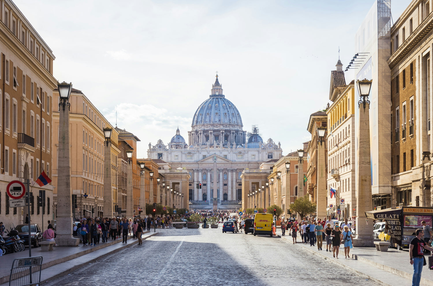 The majestic dome of St. Peter's Basilica rises at the end of a lively street in Rome, filled with pedestrians, cars, and vibrant city life. The avenue is bordered by classical buildings and stone columns, leading the eye toward the iconic structure.