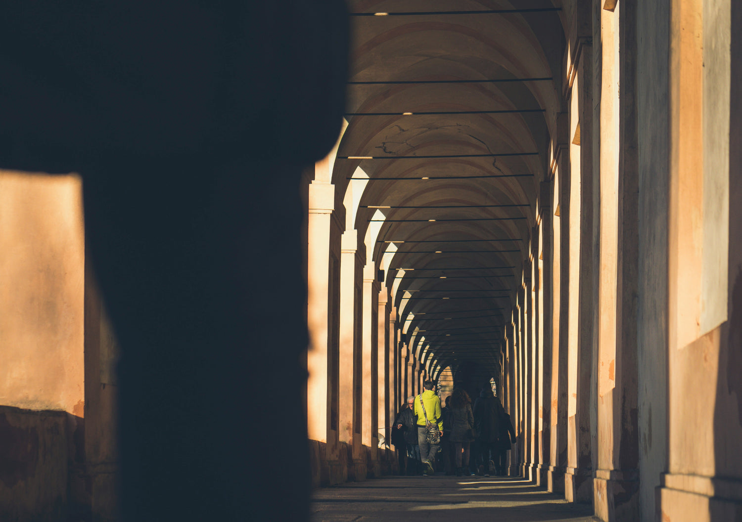 A narrow, sunlit corridor with a series of archways creates a tunnel-like effect, leading to a distant group of people. The shadows cast by the arches add a rhythmic pattern to the stone floor and walls, highlighting the architectural symmetry.