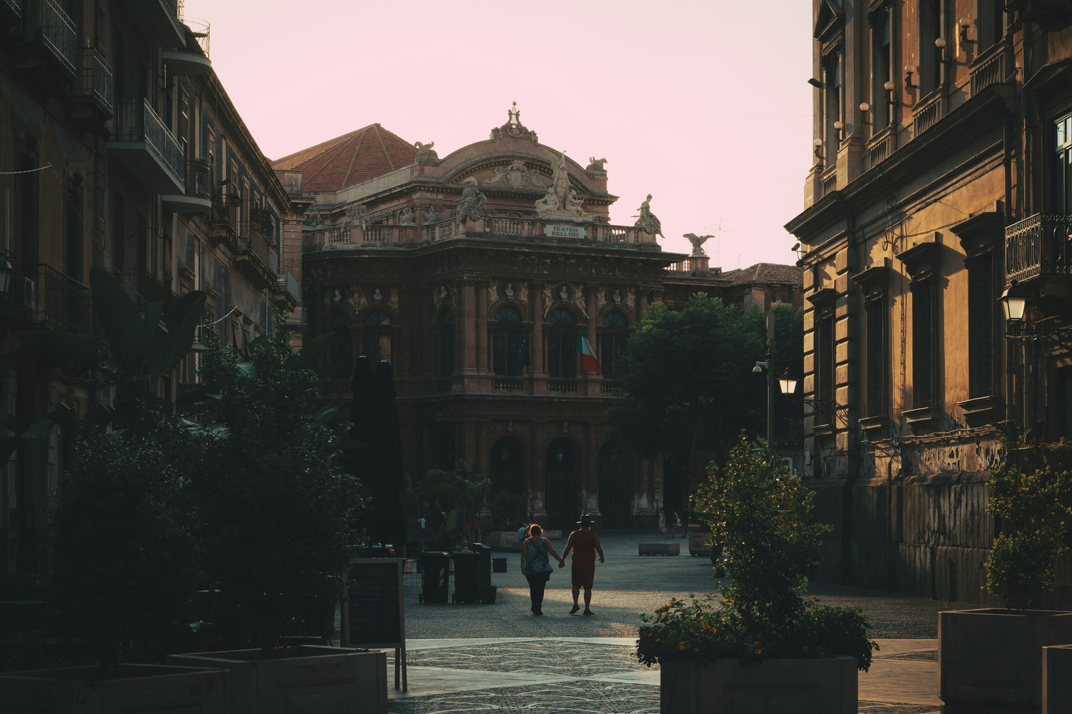 A serene evening scene shows a couple walking through a plaza toward an imposing theater building with a richly decorated facade. The soft pink sky contrasts with the dark shadows of the surrounding buildings, creating a peaceful, reflective mood