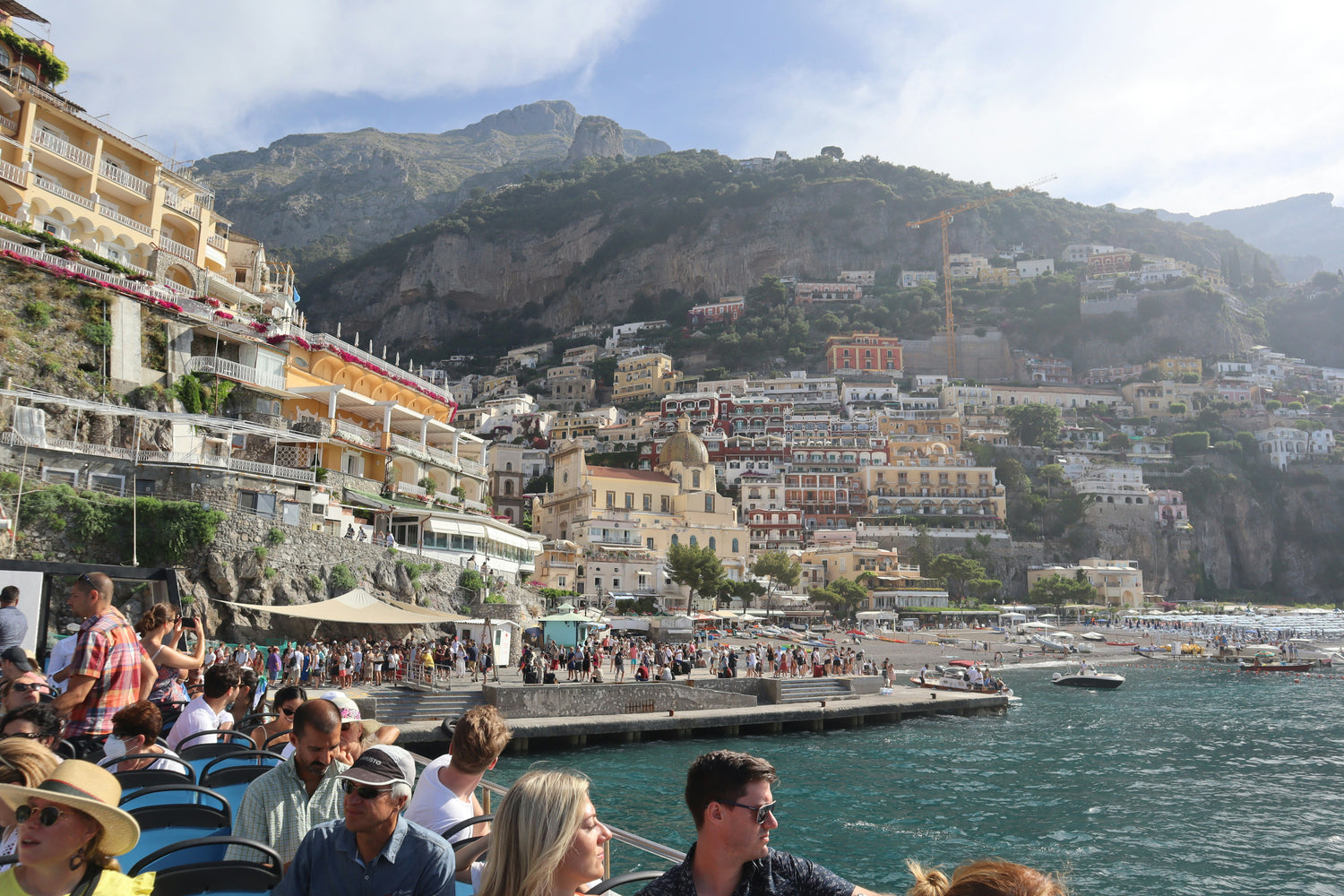 A coastal town built on steep cliffs, with colorful buildings cascading down the hillside toward a busy waterfront. The scene includes tourists seated on a boat in the foreground, enjoying the view of the picturesque town and clear blue water.