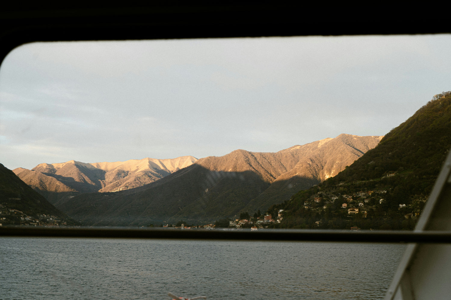 A view of sunlit mountains seen through the window of a moving vehicle or boat. The warm light highlights the rugged terrain, casting deep shadows in the valleys, while a calm lake stretches out in the foreground.