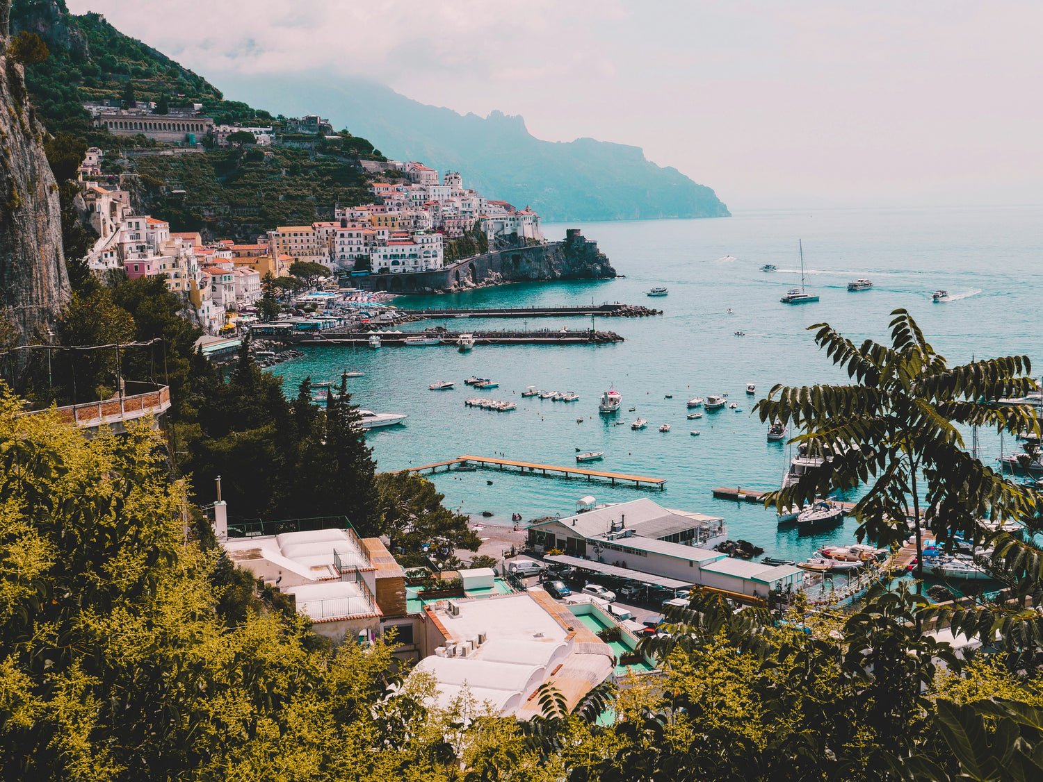 A coastal town with buildings on a hillside overlooking a bay filled with boats, set against a backdrop of mountains and cloudy sky.